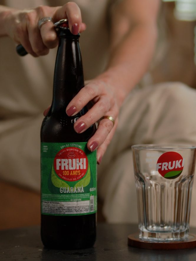 Mãos abrindo garrafa de guaraná Fruki ao lado de um copo, cena de comercial