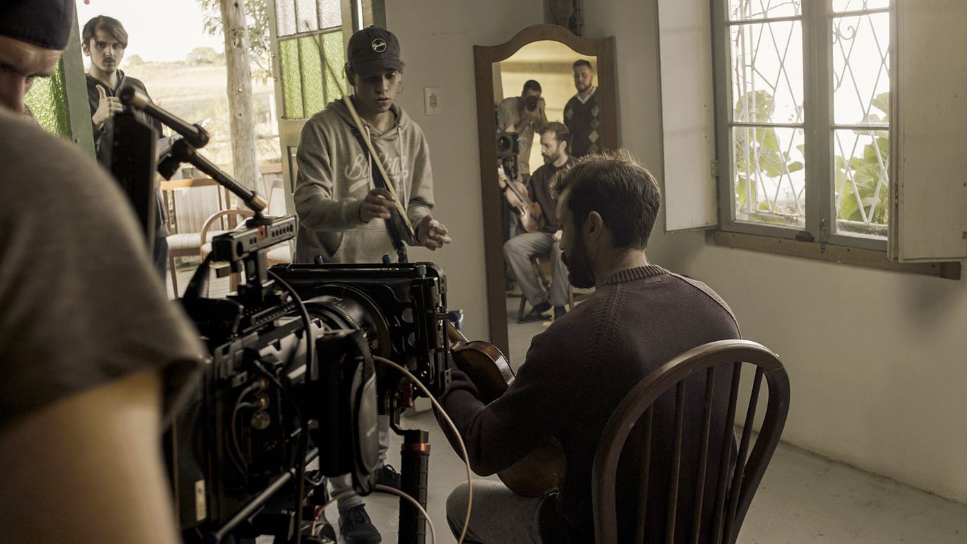 Equipe preparando cena com ator tocando violino em casa de campo, câmera de cinema em primeiro plano