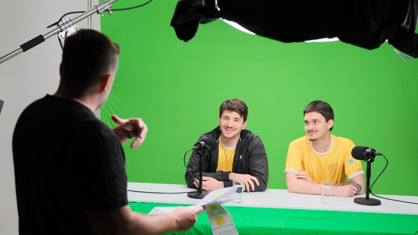Diretor orientando dois apresentadores em bancada de chroma key verde com microfones
