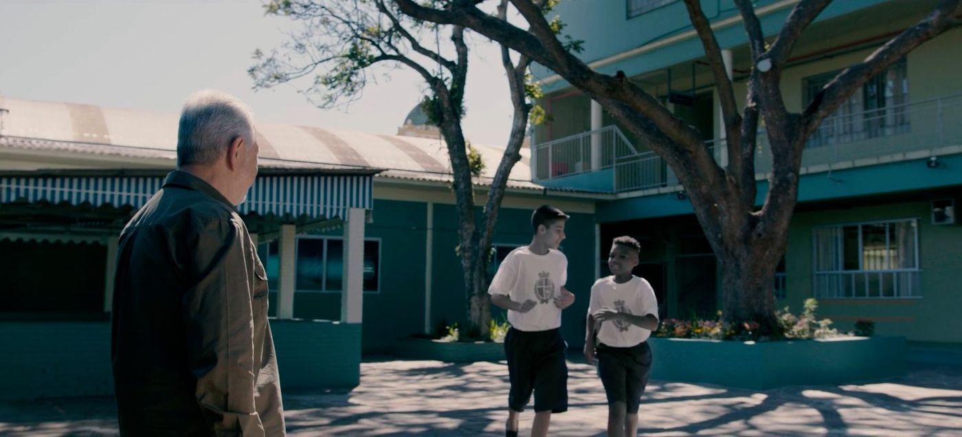 Homem idoso observa dois alunos correndo no pátio de uma escola, cena de filme institucional